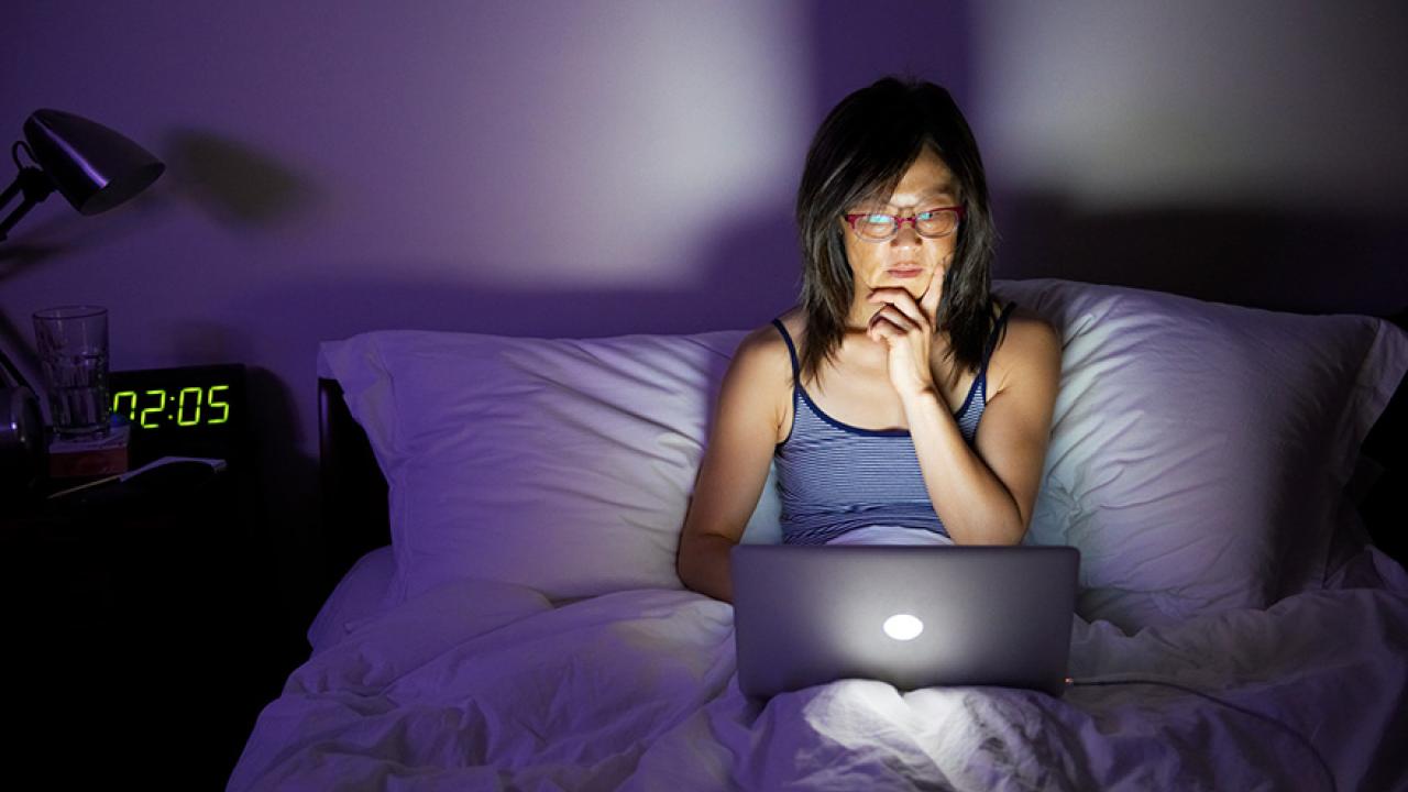 A woman sits in bed in the dark on her laptop.