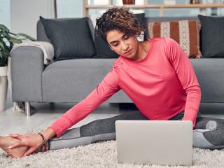 A woman does yoga in her living room while watching a laptop, placed on the floor.
